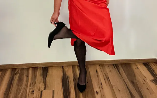 Man in a red satin skirt and black tights lifting one foot in a black high-heeled pump